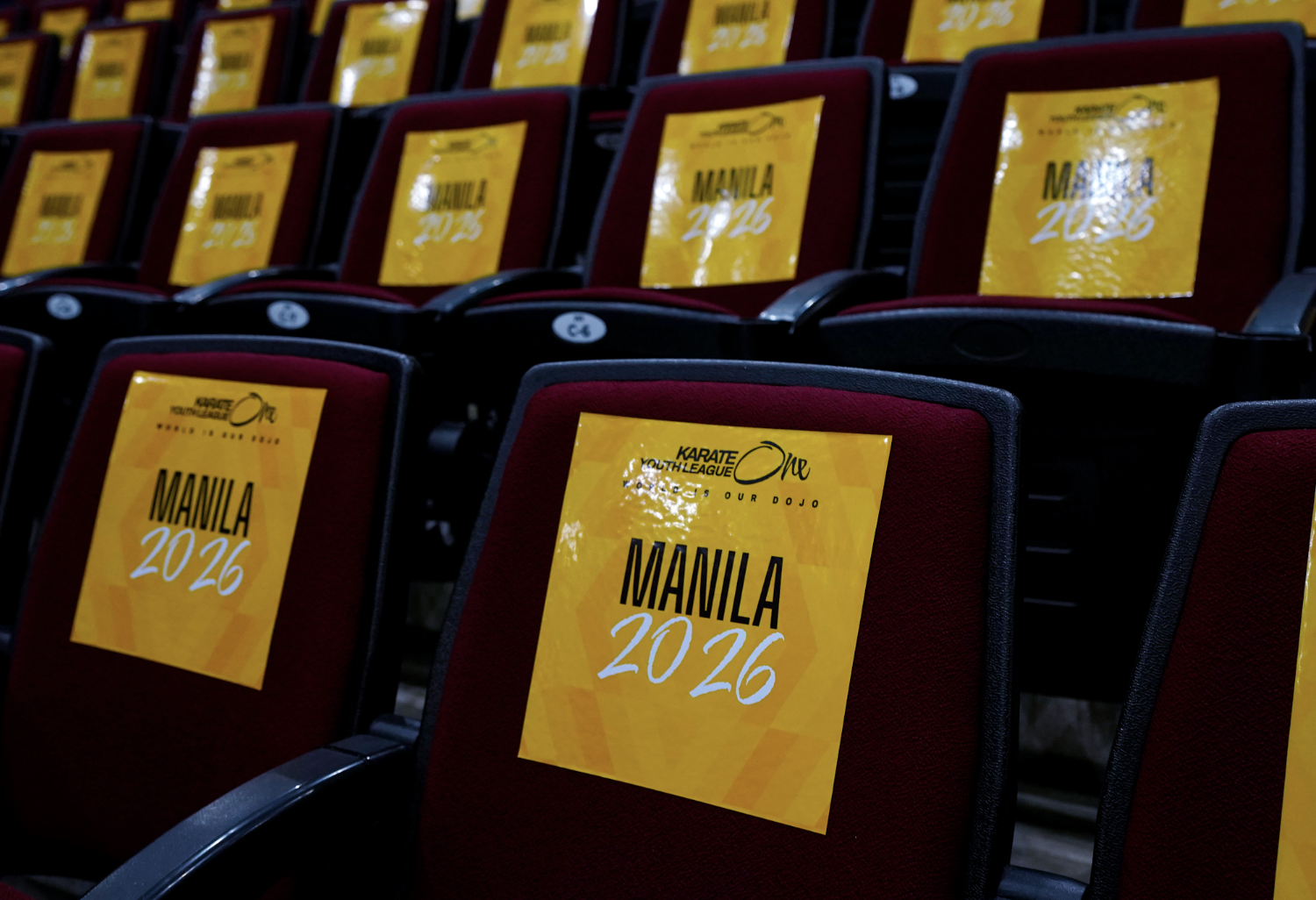 Multiple empty stadium seats with yellow signs reading 'MANILA 2026' attached to the backrests.