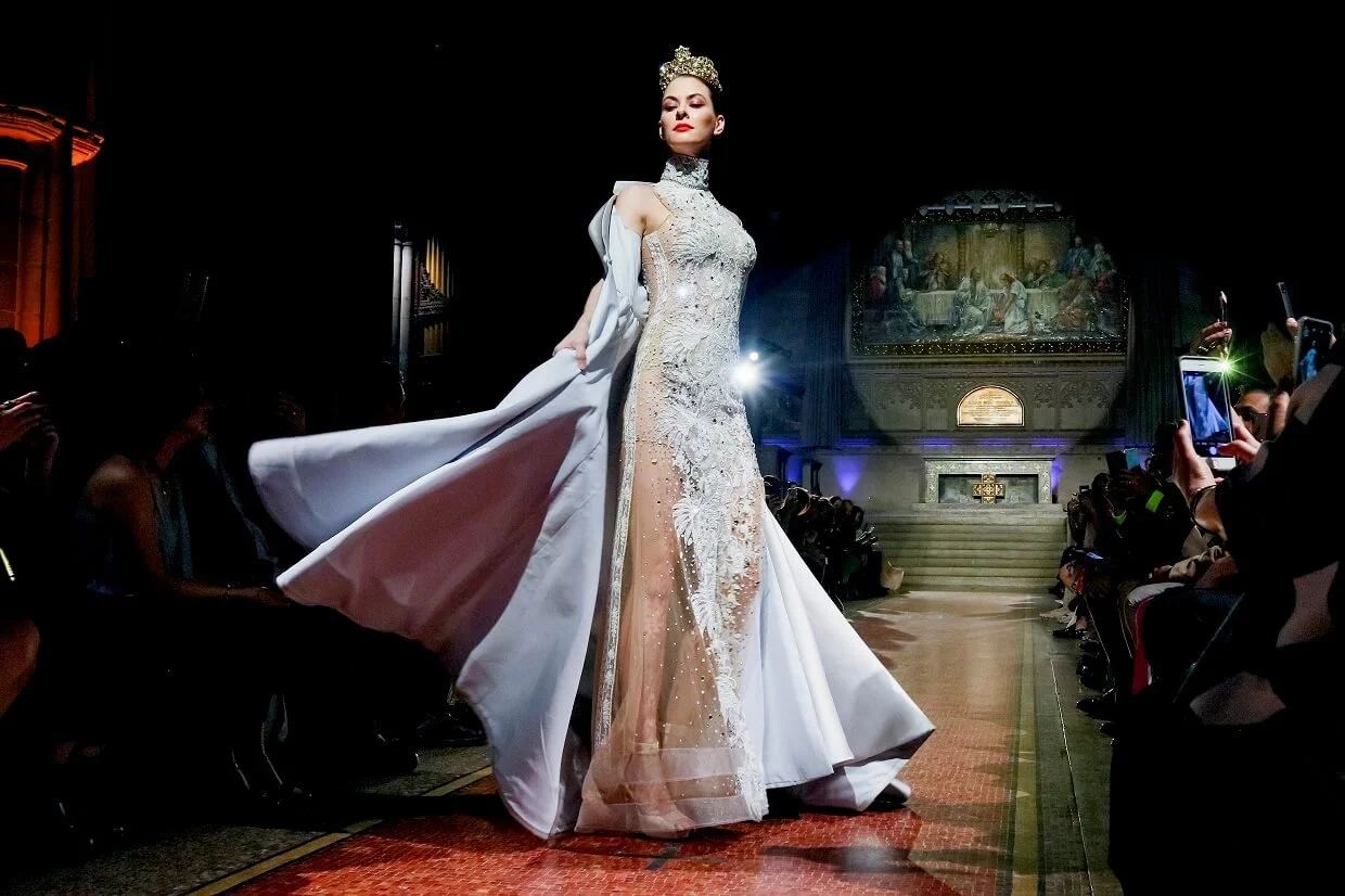A woman walking down a runway during a fashion show wearing an ornate, high-fashion gown with a crown. Audience members are taking photos on either side of the runway.
