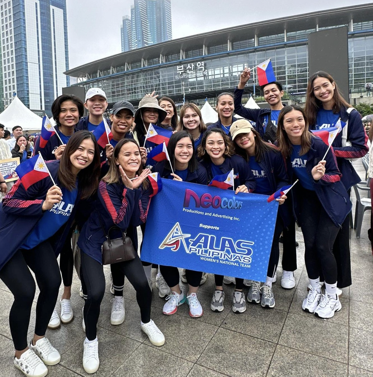 Group of women holding Filipino flags and a banner supporting Alas Pilipinas Women's National Team outside a modern building with skyscrapers in the background.