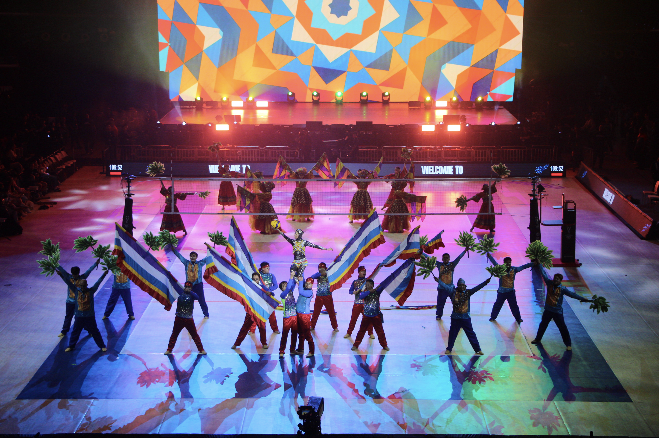 Performers in colorful costumes waving flags and holding bouquets on a brightly lit stage with a large, colorful geometric backdrop, and a digital sign that reads 'Welcome to'.
