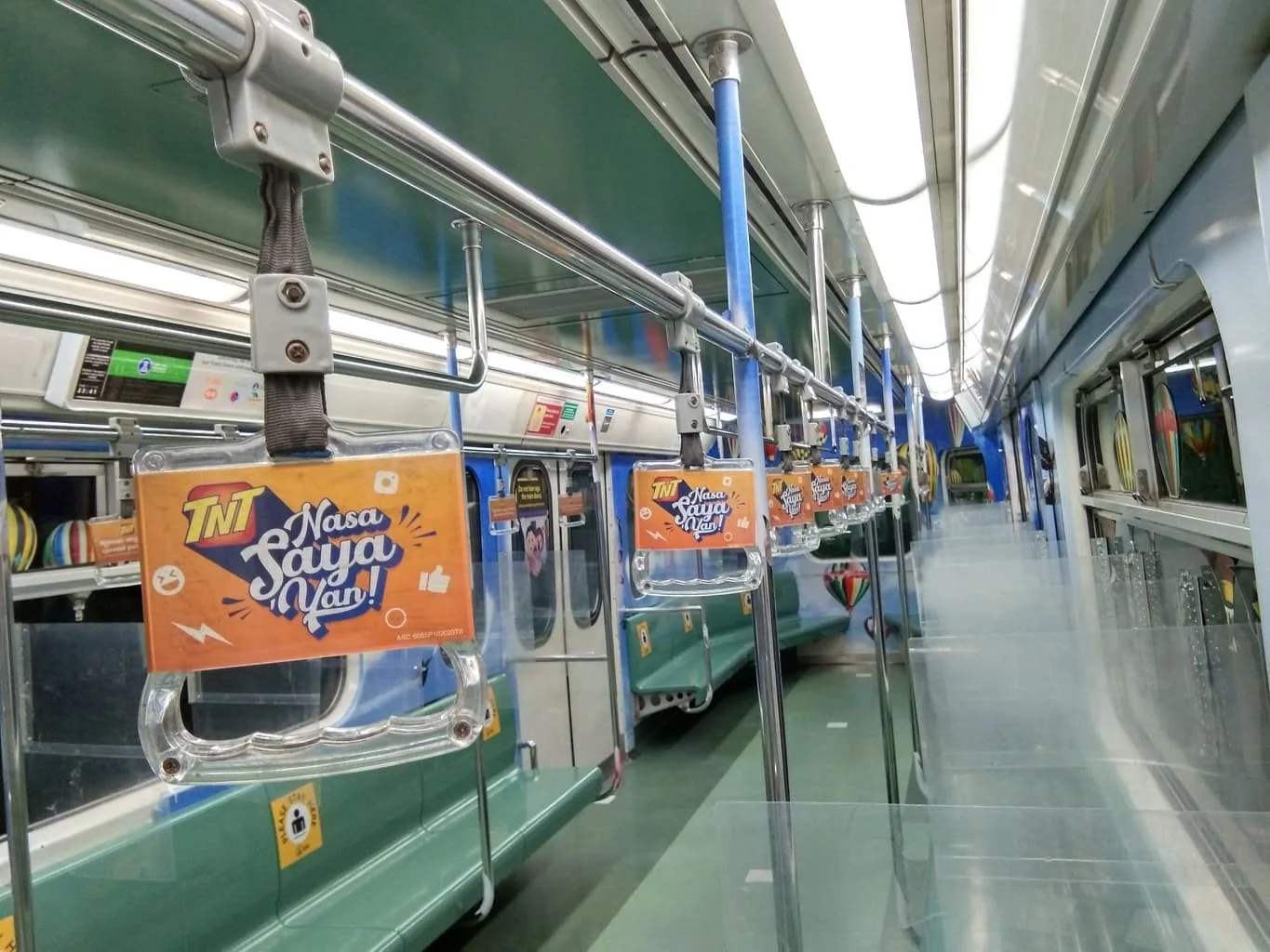 Empty subway car with green seats, some advertising signs hanging from the ceiling, and a row of windows on the right side.