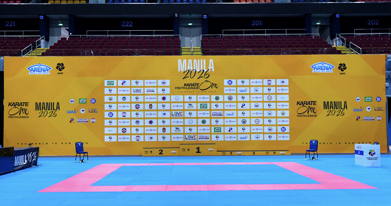 Karate competition area with a blue mat and a pink boundary, chairs on either side, and a yellow backdrop with logos and text indicating the Manila 2026 Karate Youth League tournament.