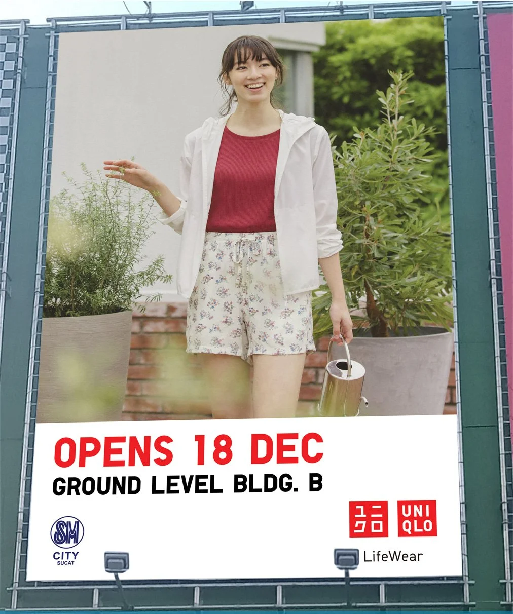 A woman standing outdoors with plants around her, smiling and holding a watering can, in front of a billboard advertising a store opening on December 18th at Ground Level Bldg. B, featuring the UNIQLO and LifeWear logos.
