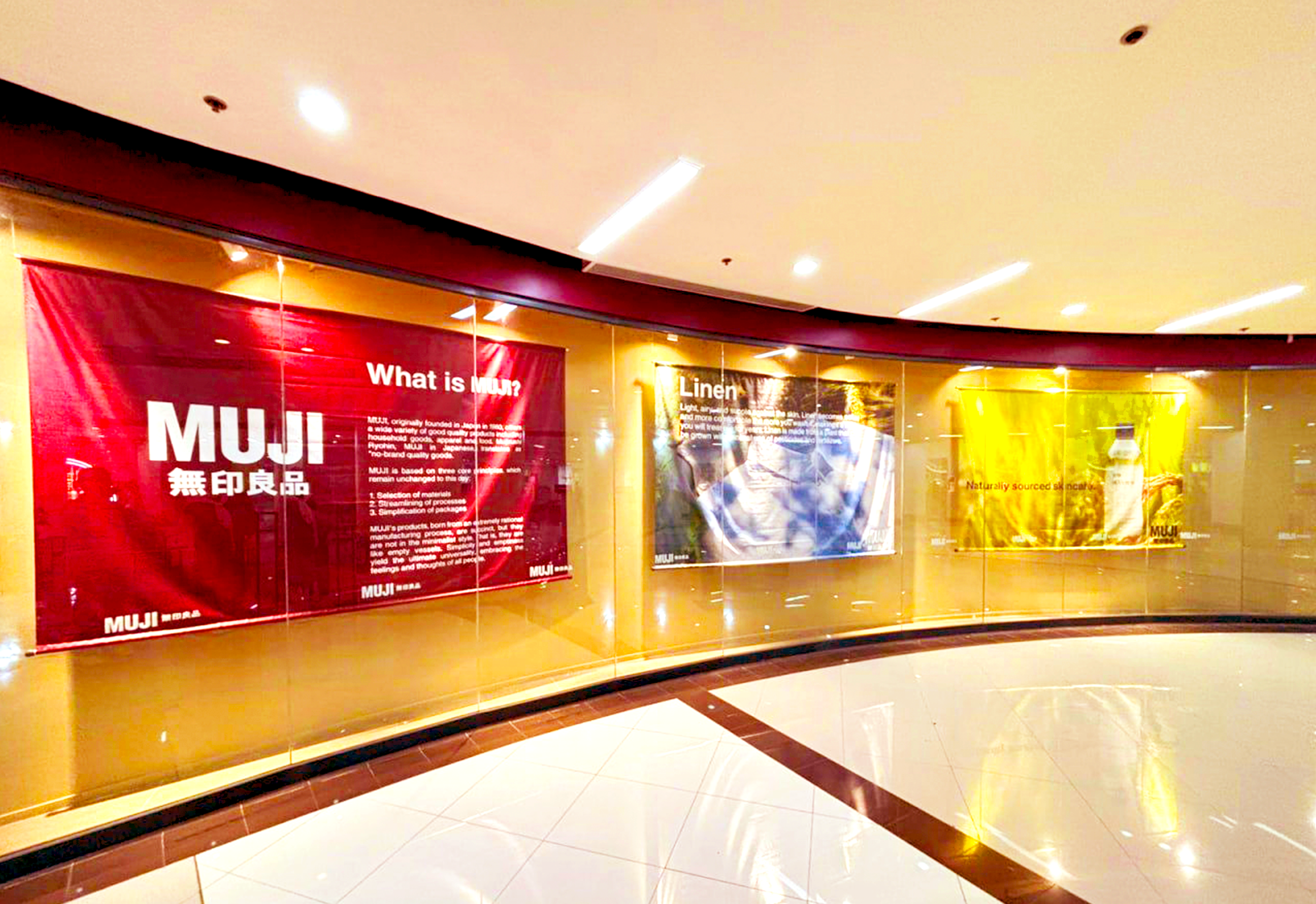 Inside view of a shopping mall with red and yellow posters promoting MUJI store. The posters contain text about MUJI and its products, displayed on a curved glass wall with a polished tiled floor.