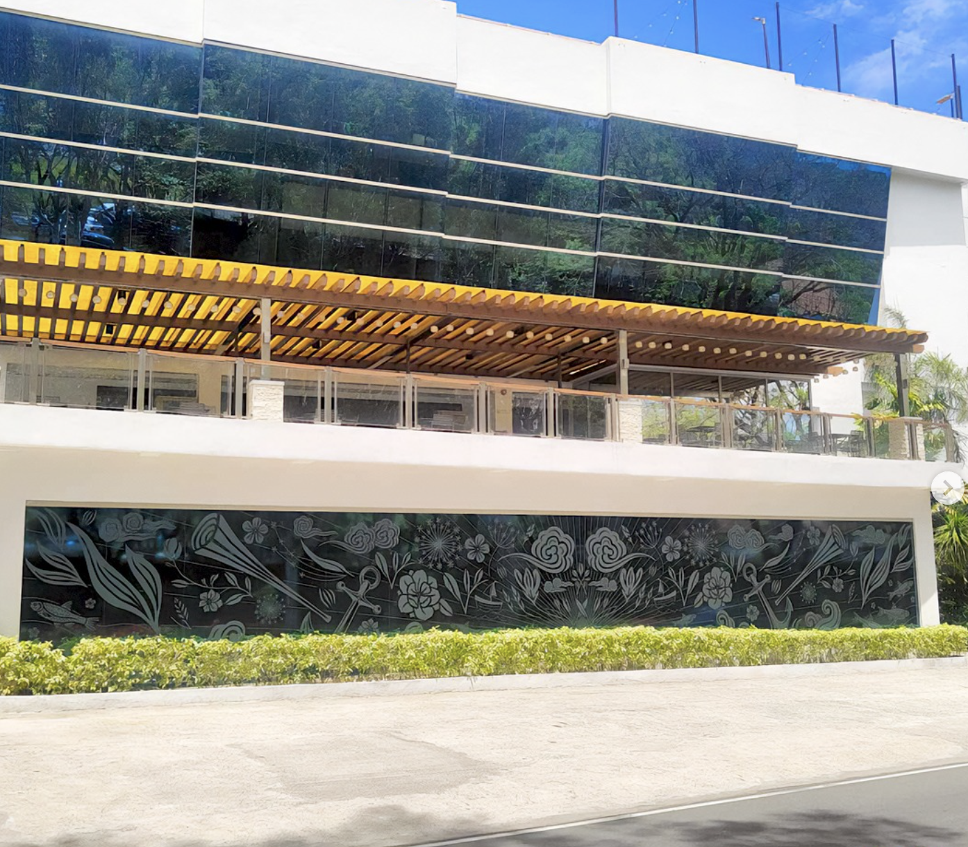 Exterior view of a modern building with glass windows, a decorative mural on the lower level, and a covered outdoor patio with a yellow awning and glass railings.