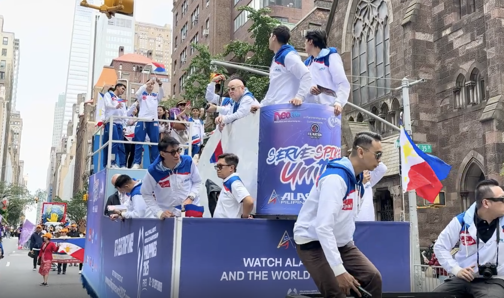 People participating in a parade on a city street, with some waving flags and dressed in white and blue. A float with a banner that reads 'Serve, Spark, United' and other text is visible, with individuals walking alongside and around it, including a 