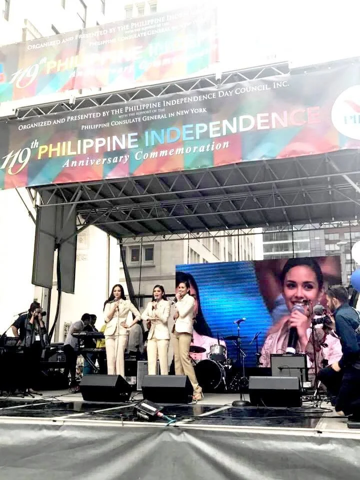 Stage with three women singing into microphones, a large screen displaying a smiling woman, and a banner celebrating the 119th Philippine Independence Day in New York City.