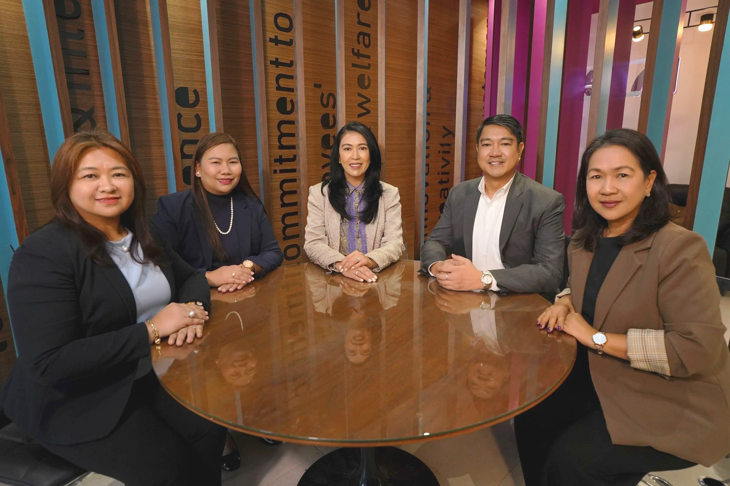 Six diverse professionals sitting around a round wooden table in a modern office with wooden and colorful wall panels. They are dressed in business attire, smiling, and posing for the photo.