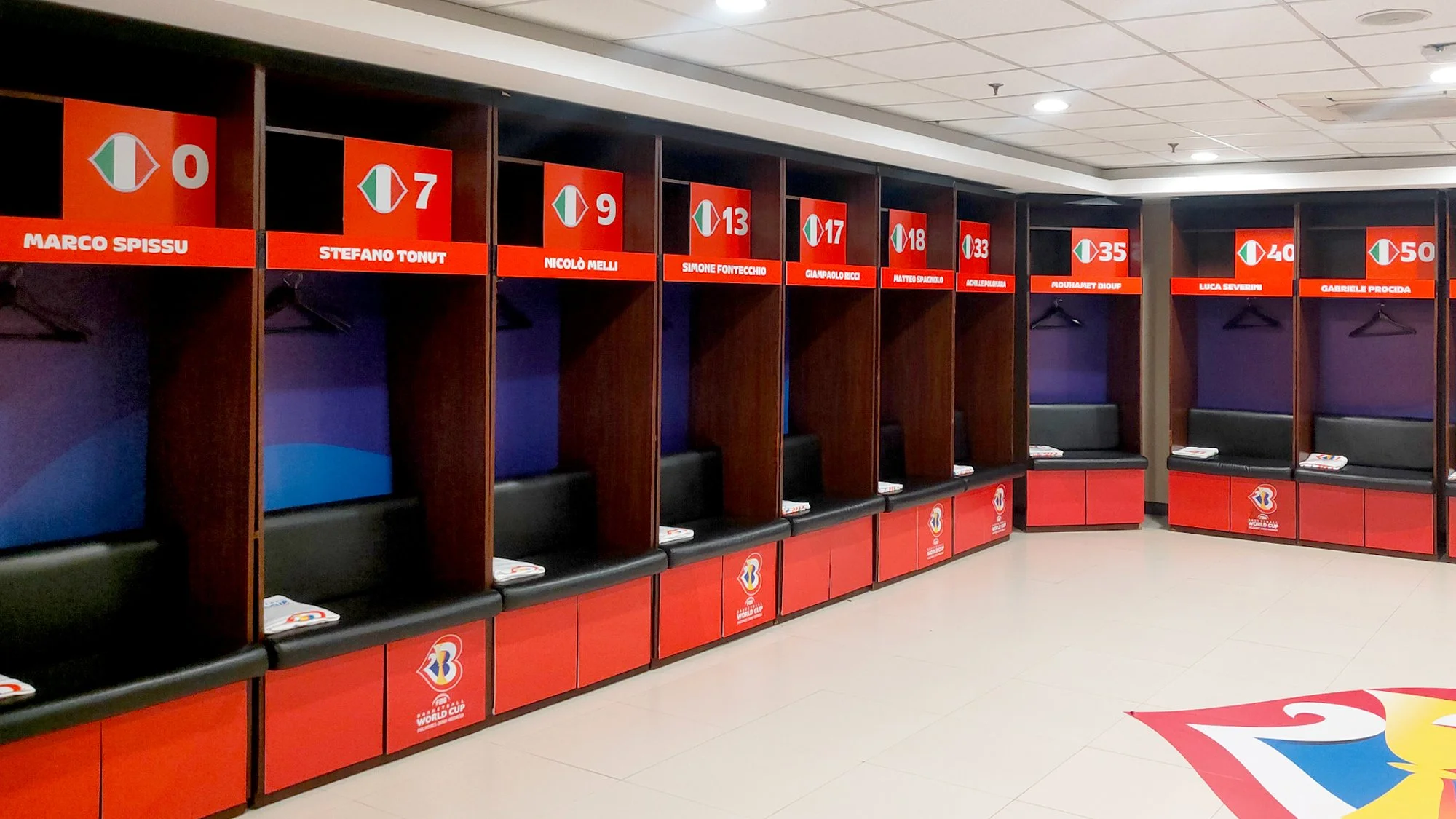 Empty locker room stalls with name tags and numbers, featuring benches and shelving. A logo related to the World Cup is visible on some of the lockers, and there is a colorful emblem on the floor.