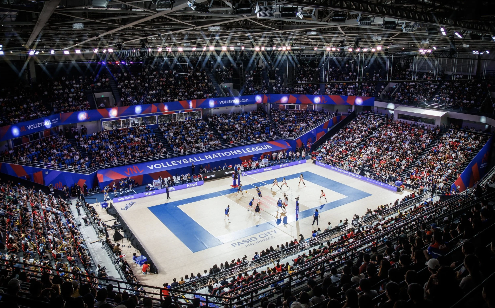 Indoor volleyball arena filled with spectators, players on the court, and banners displaying 'Volleyball Nations League.'