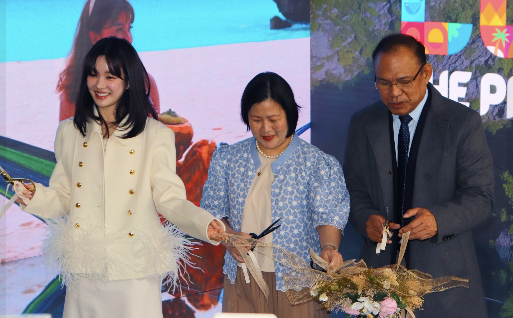 Three people in formal attire participating in a ribbon-cutting ceremony, with a large digital screen displaying a beach scene and colorful graphics in the background.