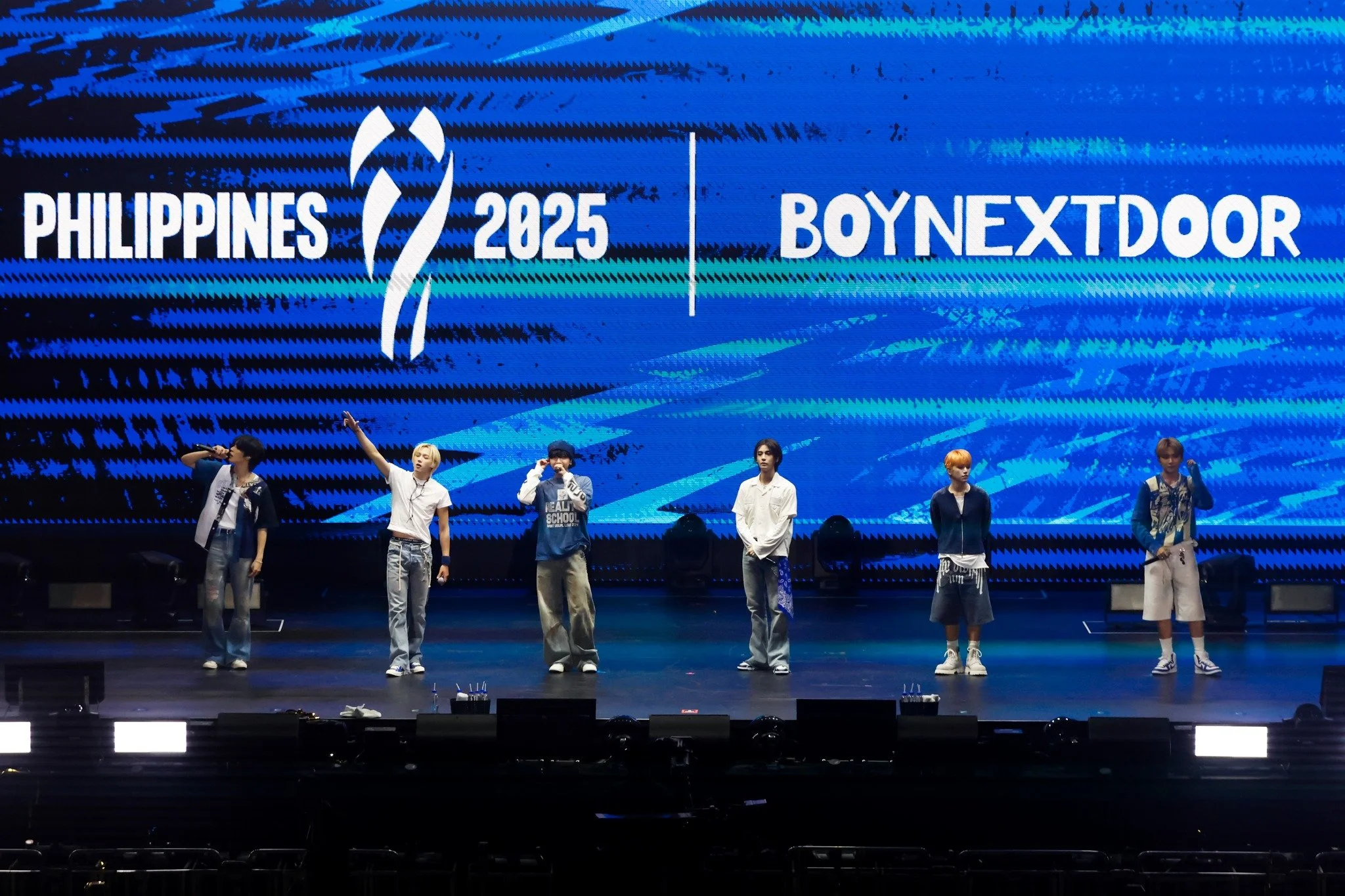 Six young men performing on stage in front of a large digital screen displaying the words "PHILIPPINES 2025 BOYNEXTDOOR".