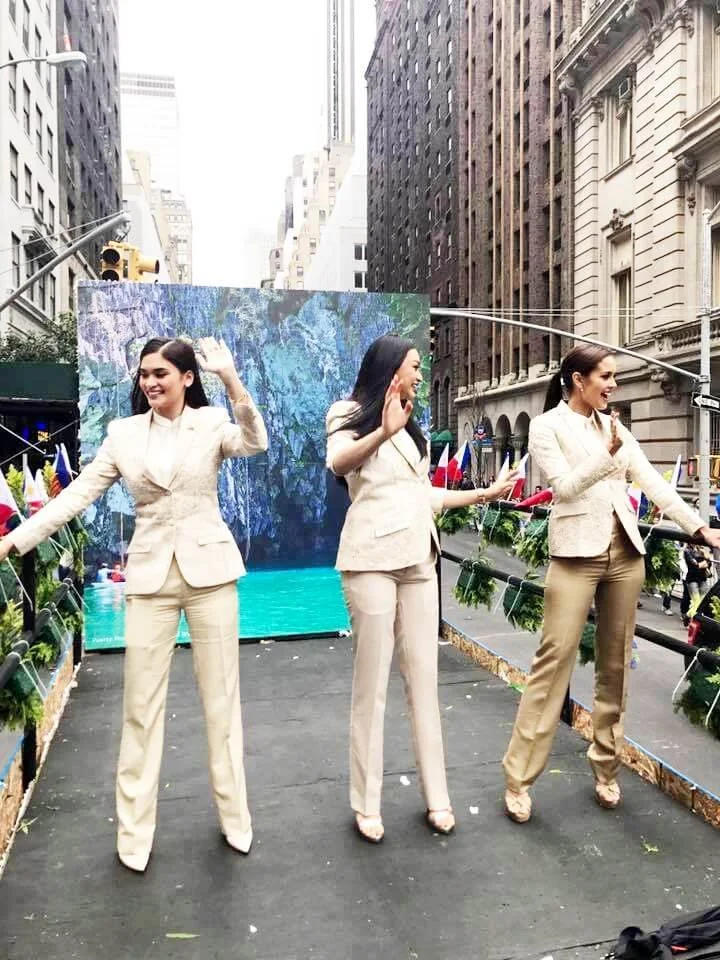 Three women in beige suits standing on a city street platform, smiling and waving at the camera, with tall buildings in the background and flags on each side of the platform.