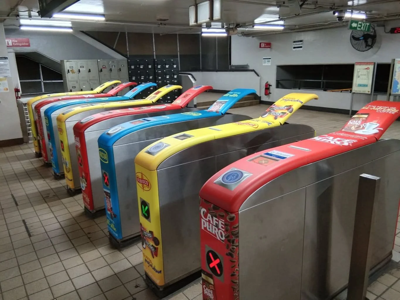 Multiple turnstiles in a subway station entrance with colorful advertising panels, including red, yellow, and blue, and lockers in the background.