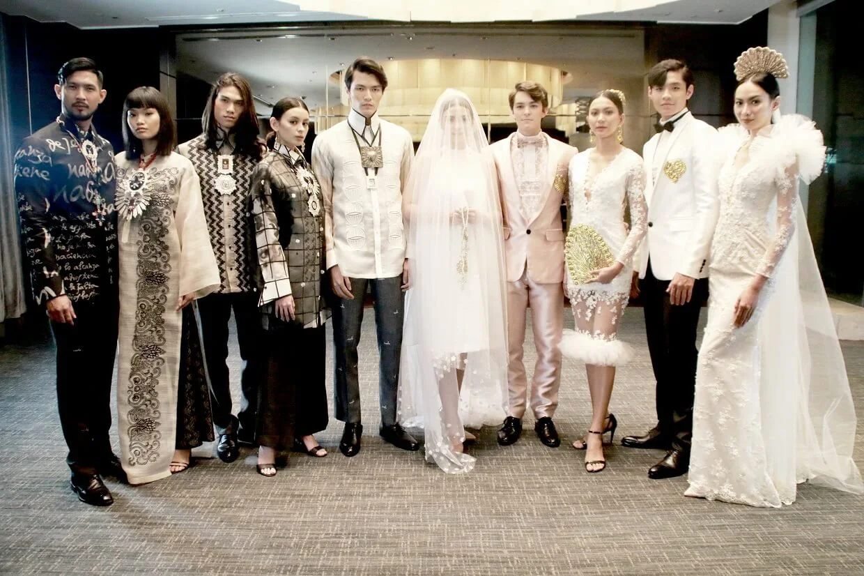 A group of people dressed in formal and traditional attire, standing in a line at a wedding or special event inside a modern venue. The woman in the center is wearing a white wedding dress with a veil, surrounded by men and women in elegant clothing.
