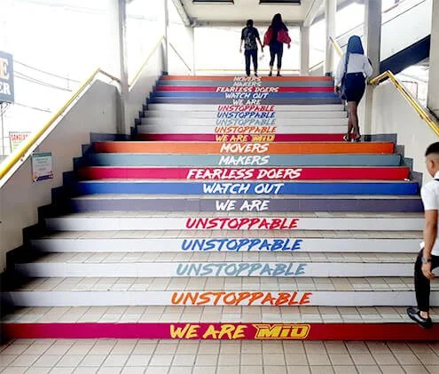 Colorful stairs with motivational phrases including 'Fearless Doers,' 'Watch Out,' 'We Are,' and 'Unstoppable,' leading up to an outdoor walkway with three people walking.
