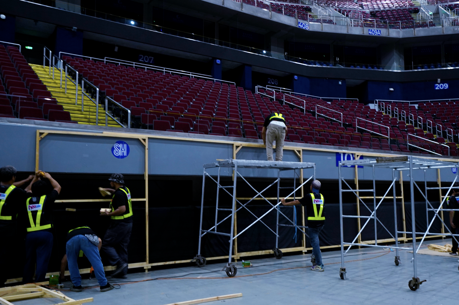 Construction workers assembling a stage in an arena with red seats and multiple levels.