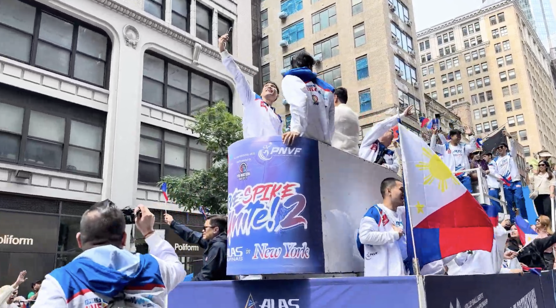 A parade float with Filipino athletes celebrating, surrounded by a crowd on a city street with tall buildings. The float displays banners with the text 'SPIKE to win!' and  'AS in New York'. Some individuals are waving Filipino flags and wearing team