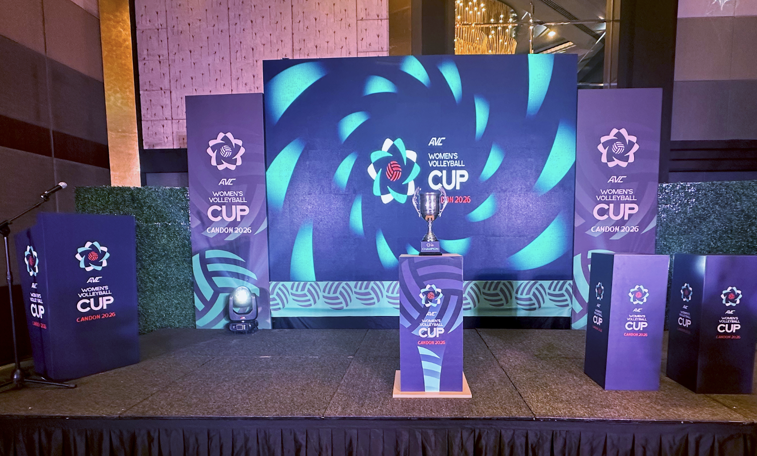 Stage set for the AVC Women's Volleyball Cup 2026, featuring a large trophy on a podium, several banners with the event logo, and a backdrop with swirling blue patterns.