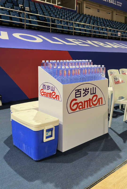 A display table with Ganten bottled water in a sports arena, with a white cooler and chairs nearby, and a large blue banner with white text in the background.