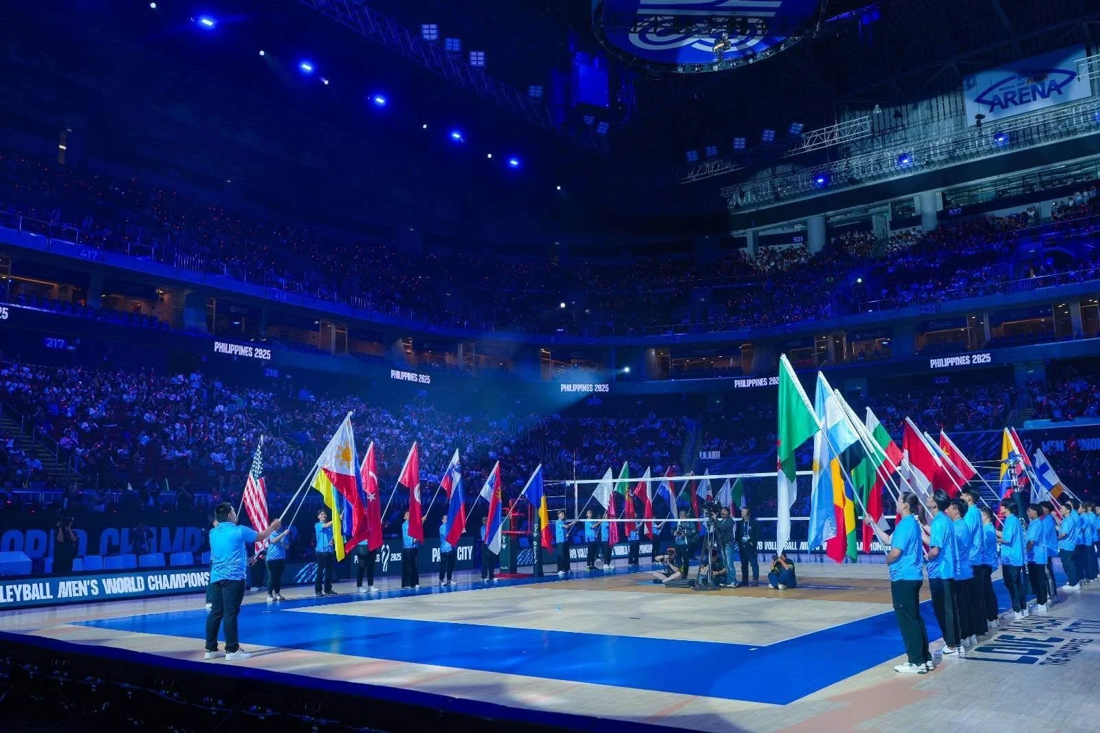 An indoor volleyball court during an international event, with athletes holding flags of different countries lined up along the court, and spectators filling the arena.