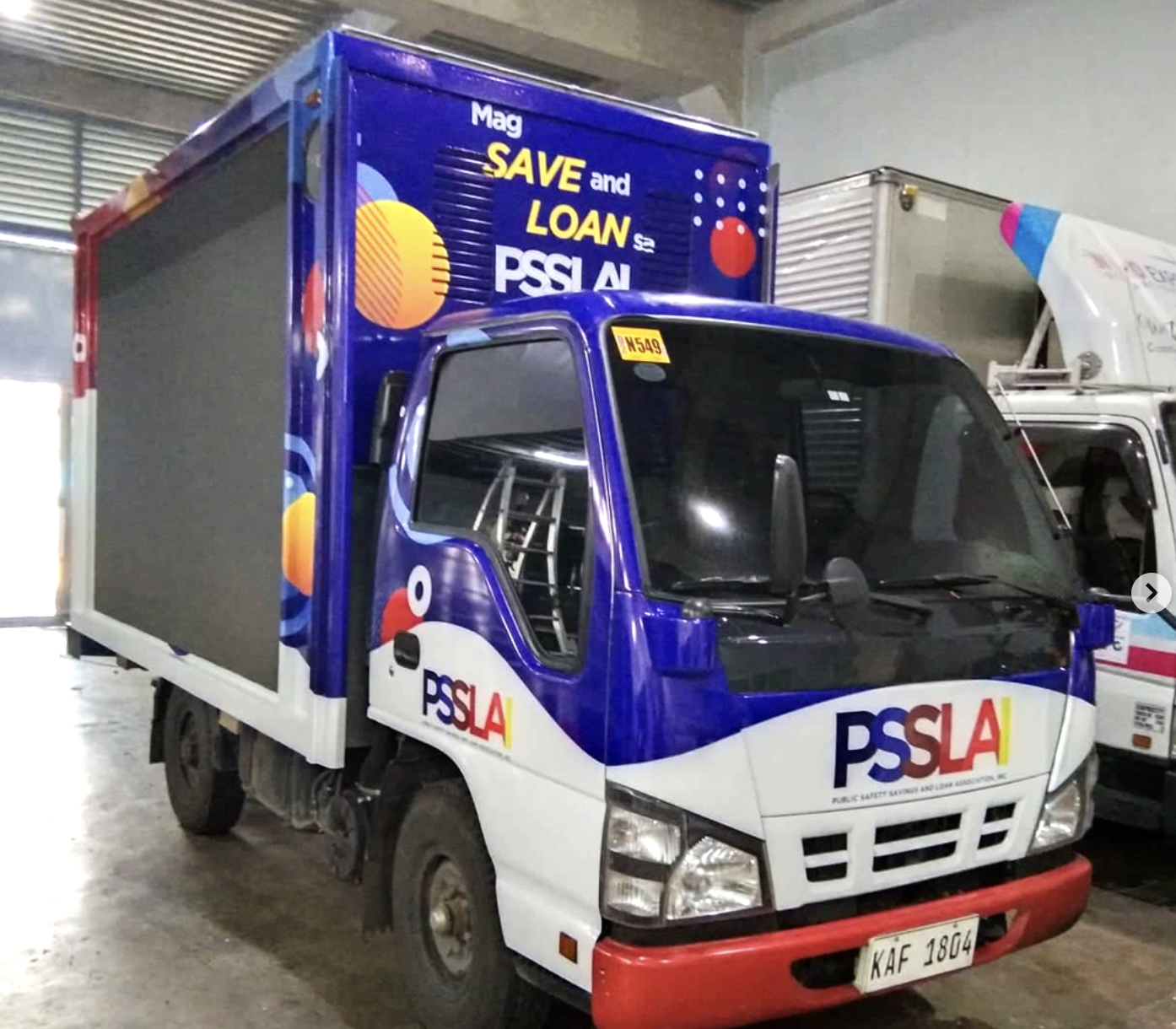 A small delivery truck with a blue, white, and red color scheme, displaying the logo 'PSSLA' and promotional text about saving and loans, parked inside a warehouse or garage.