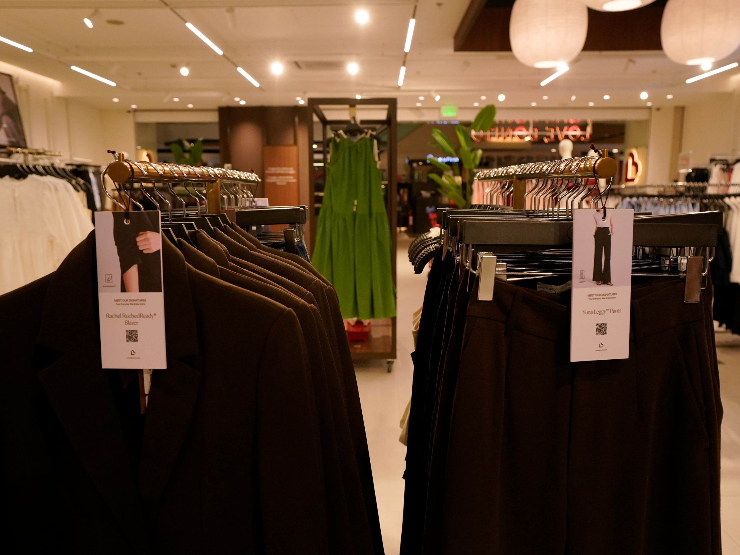 Clothing racks in a retail store with garments and price tags, with a green dress in the background and store lighting overhead.