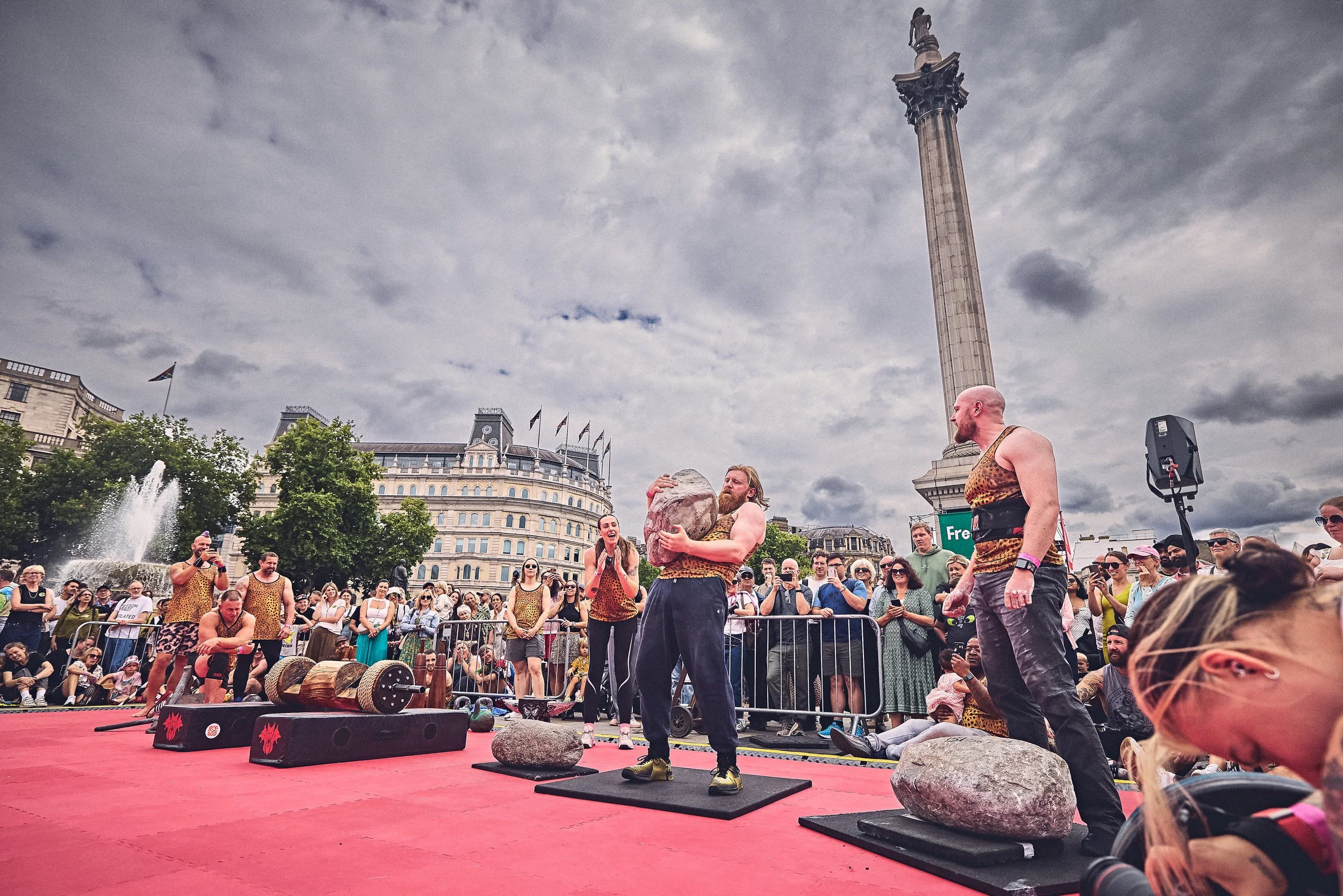 Women in animal print clothing participating in a strength competition, lifting heavy stones outdoors on a red mat, with a crowd watching in front of historic buildings and a tall monument, under cloudy skies.