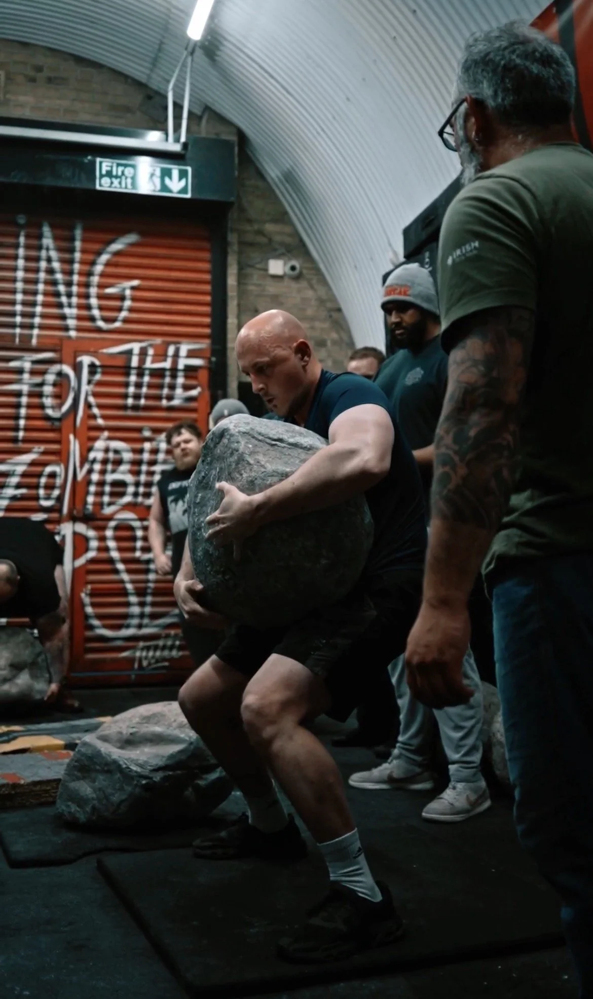 A man participating in a strongman competition, lifting a large rock while others watch in an indoor gym with graffiti art on the wall.