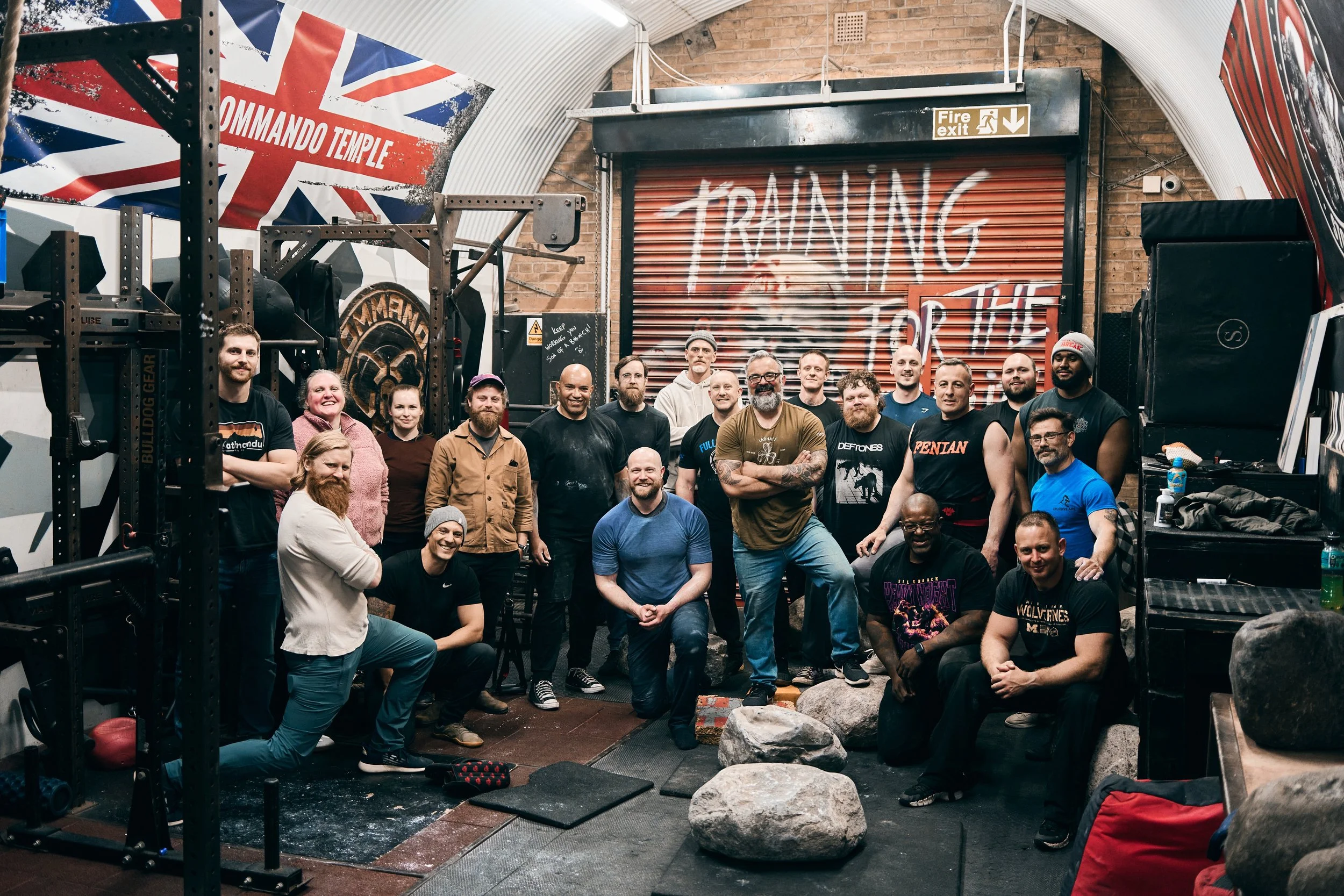 A group of people posing inside a gym with gym equipment, a large Union Jack flag with the words 'COMMANDO TEMPLE', and graffiti on a metal door that says 'TRAINING'.