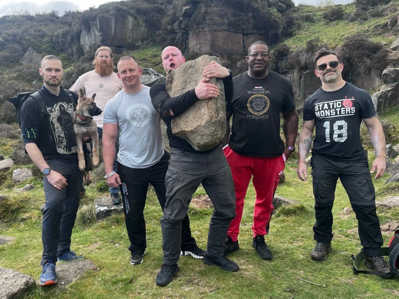 A group of six men, one holding a dog, posing outdoors on a grassy area with rocks and a rocky hillside in the background. One man is holding a large rock over his shoulder, and another is wearing a 'Black Panther Wakanda Forever' t-shirt. They are standing close together, some smiling and some making expressive faces.
