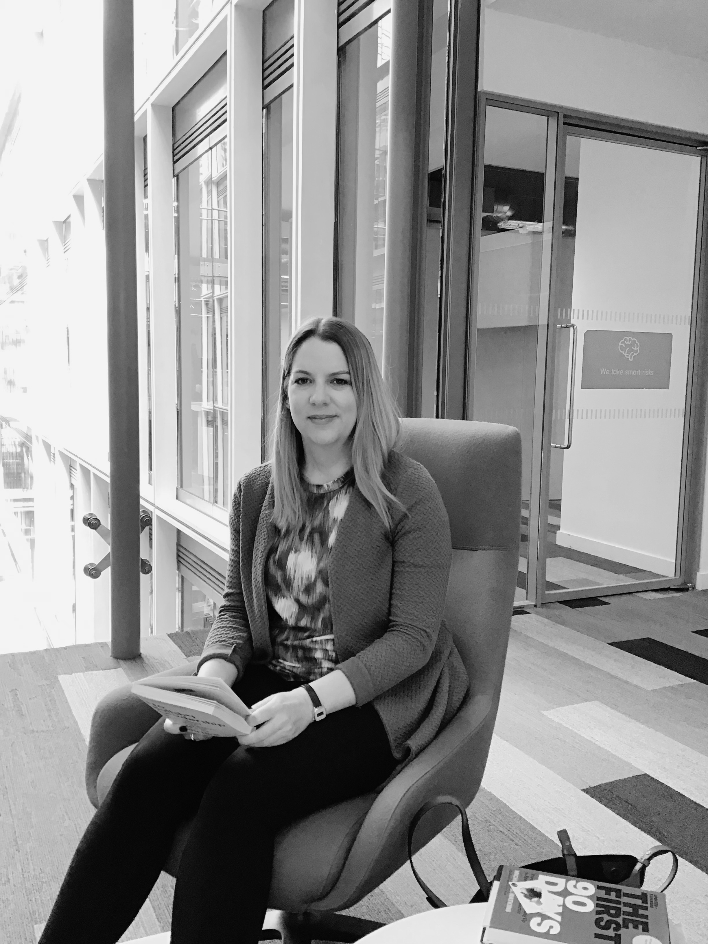 Black and white photograph of a woman sitting in a chair with a glass office background behind her.