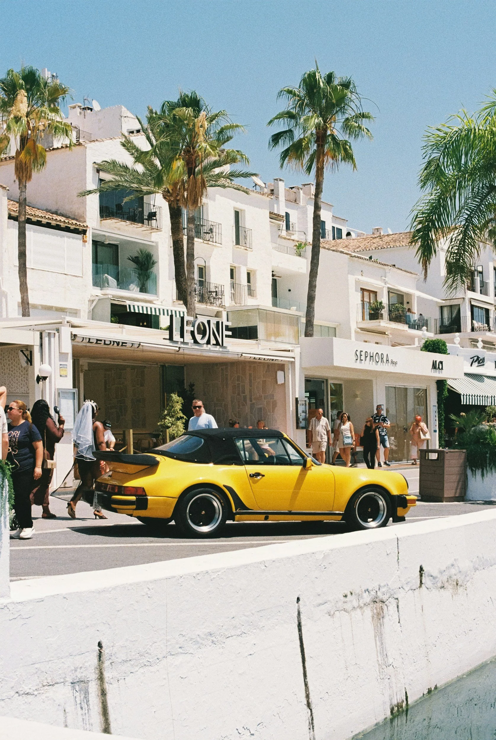 A yellow sports car parked on a city street in front of white buildings with balconies and palm trees, several pedestrians walking by under a clear blue sky.