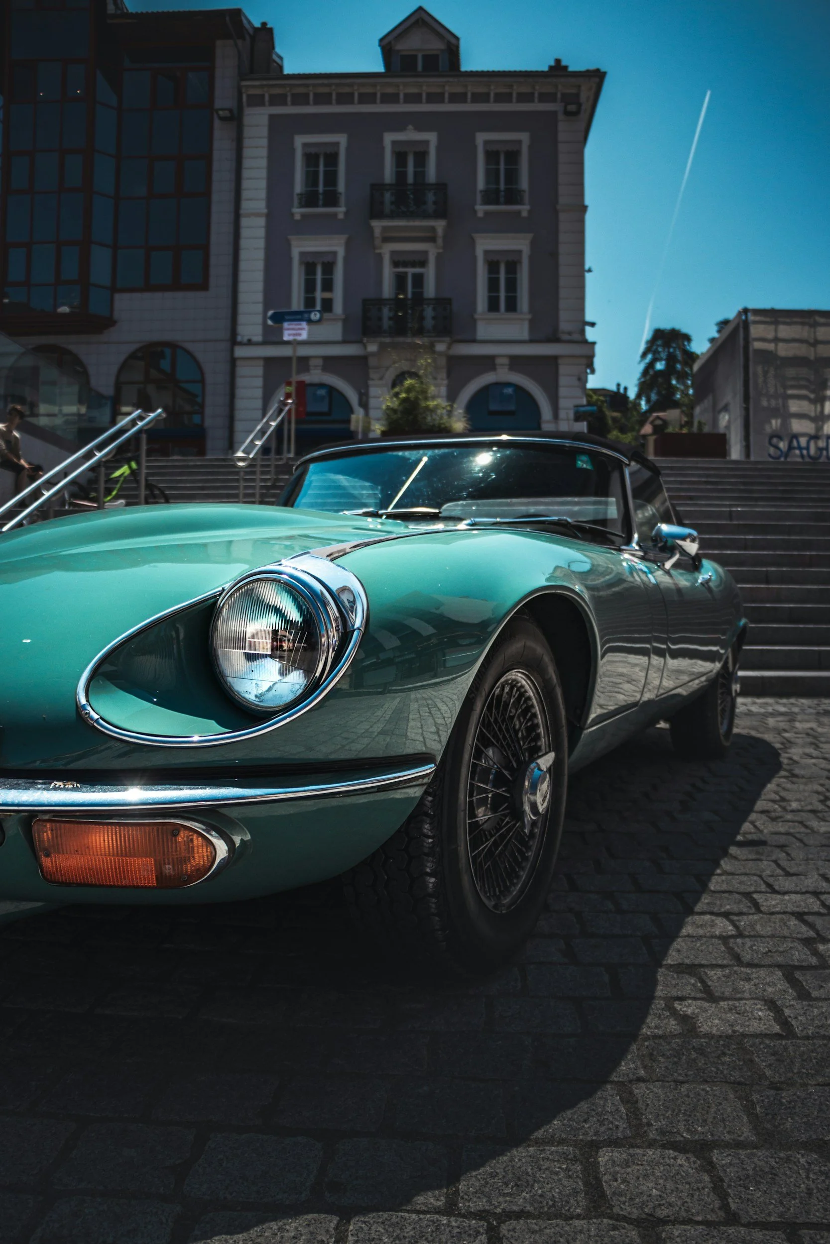 A vintage mint green sports car parked on a cobblestone street in front of stairs leading to a building with classical architecture and large windows.