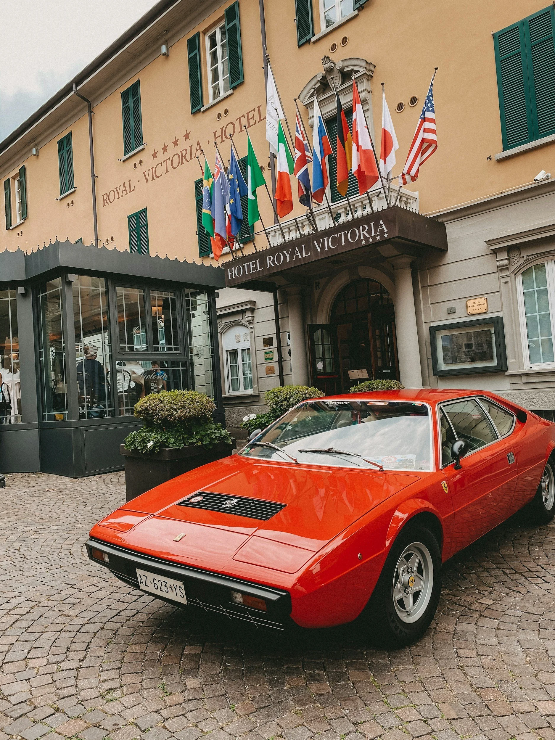 A red Ferrari sports car parked on a cobblestone street in front of a hotel with multiple flags flying above the entrance. The hotel has a beige exterior, green shutters, and a sign reading "Hotel Royal Victoria".