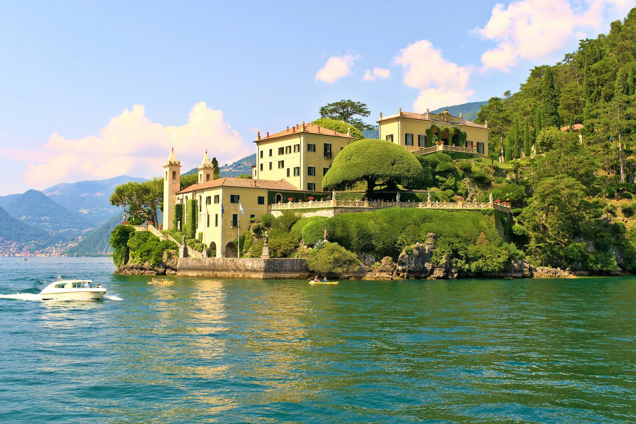 A lakeside scene with a large yellow villa on a hill surrounded by lush green trees, with mountains in the background. There are boats on the water, and the sky is blue with scattered clouds.