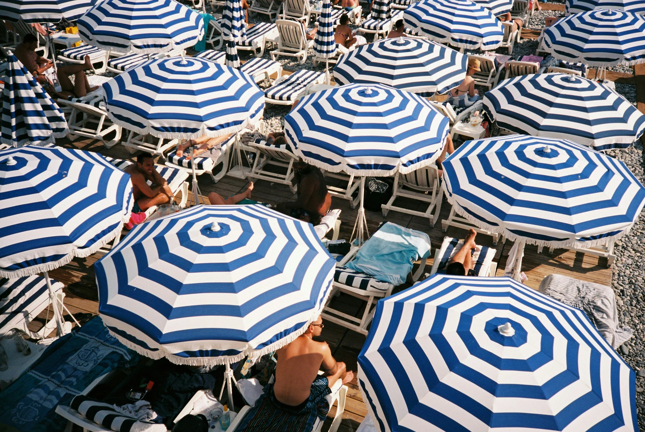 Beach scene with numerous blue and white striped umbrellas and people relaxing on lounge chairs and towels.