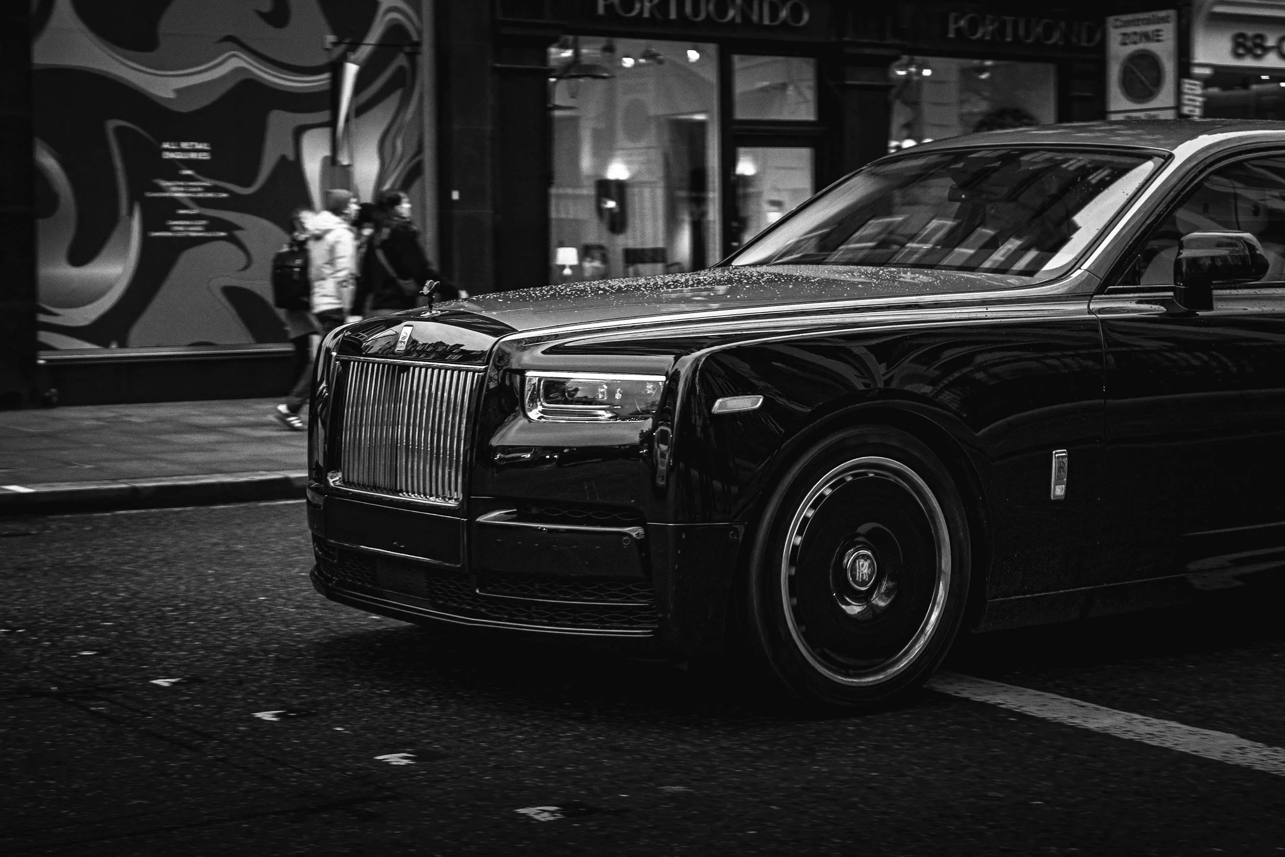 Black luxury car in motion on a city street with pedestrians passing by in the background.