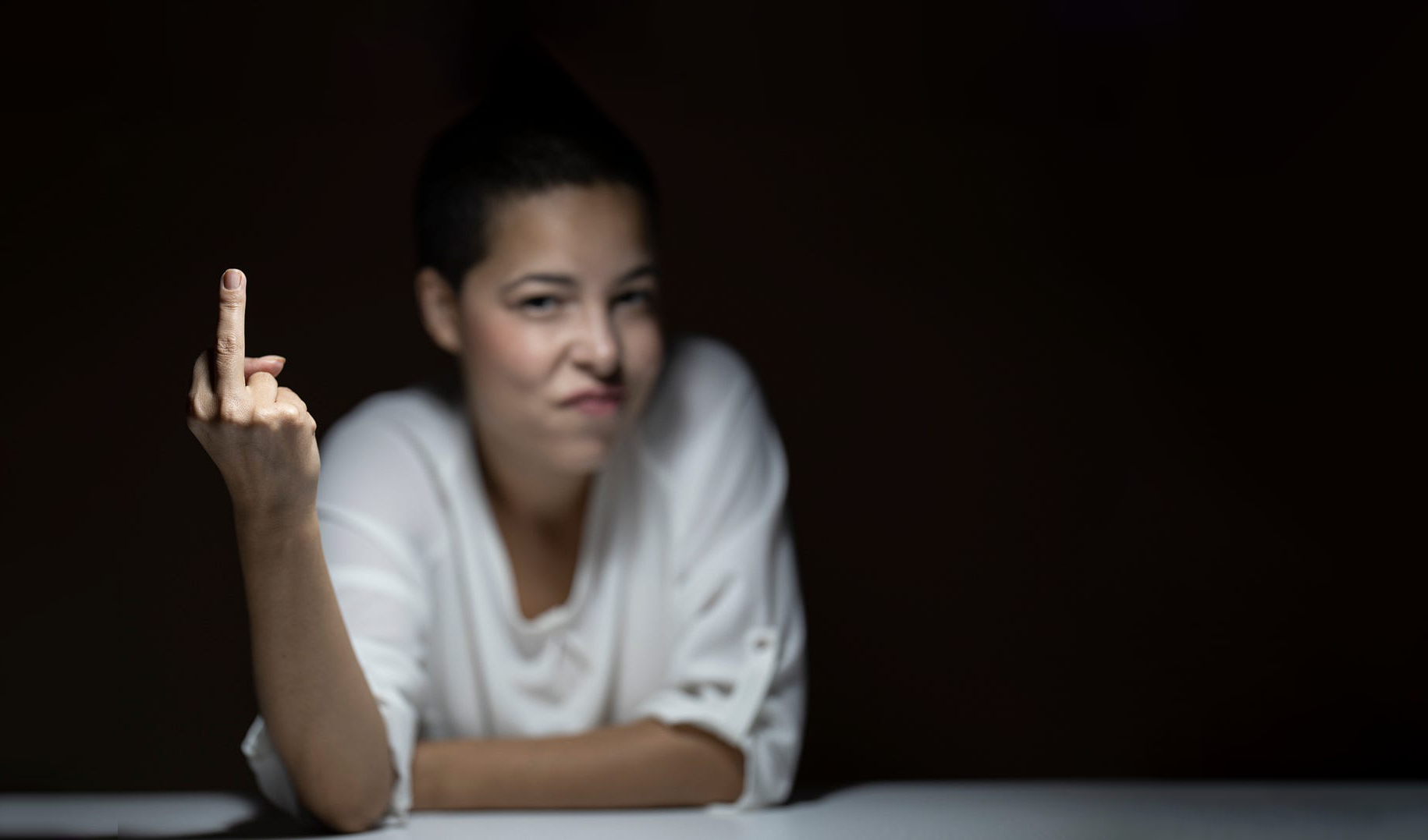 Woman with short dark hair making a disapproving or annoyed facial expression, raising her middle finger, against a dark background.