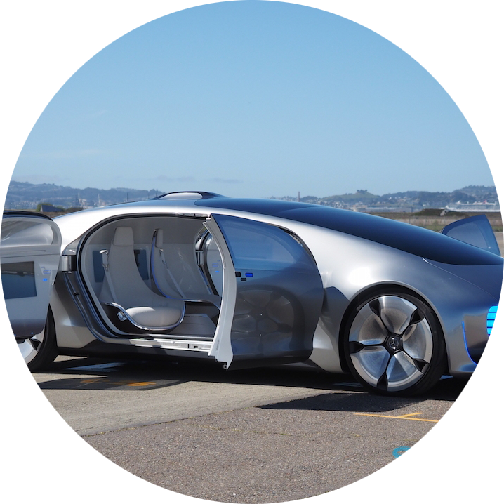 Futuristic silver concept car with open passenger side door, parked outdoors on a paved surface with a blue sky and distant landscape in the background.