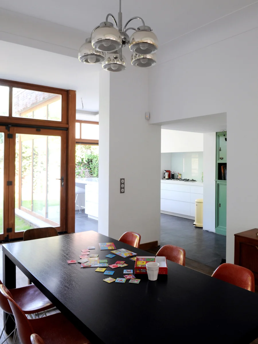 A dining room table with scattered board game pieces and a white cup, with a view of a bright kitchen and wooden and glass door leading to outside.