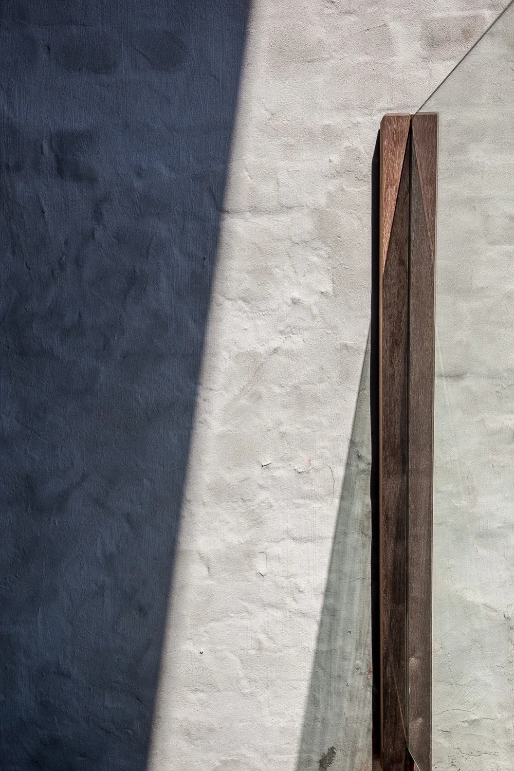 Close-up of a textured wall with a vertical wooden sculpture and a thin wire running from the top of the sculpture to outside the frame, casting shadow on the wall.