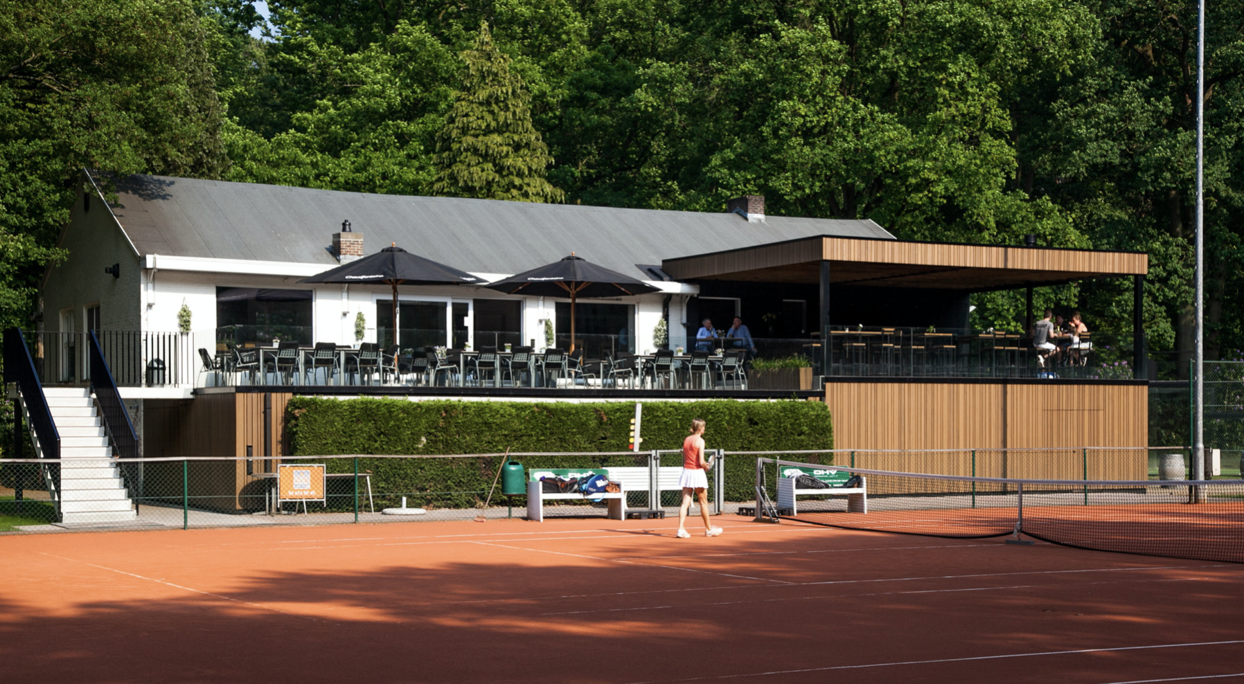 Exterior view of a two-story clubhouse with integrated outdoor seating and a contemporary architectural design by Tom Naeyaert Architecture.