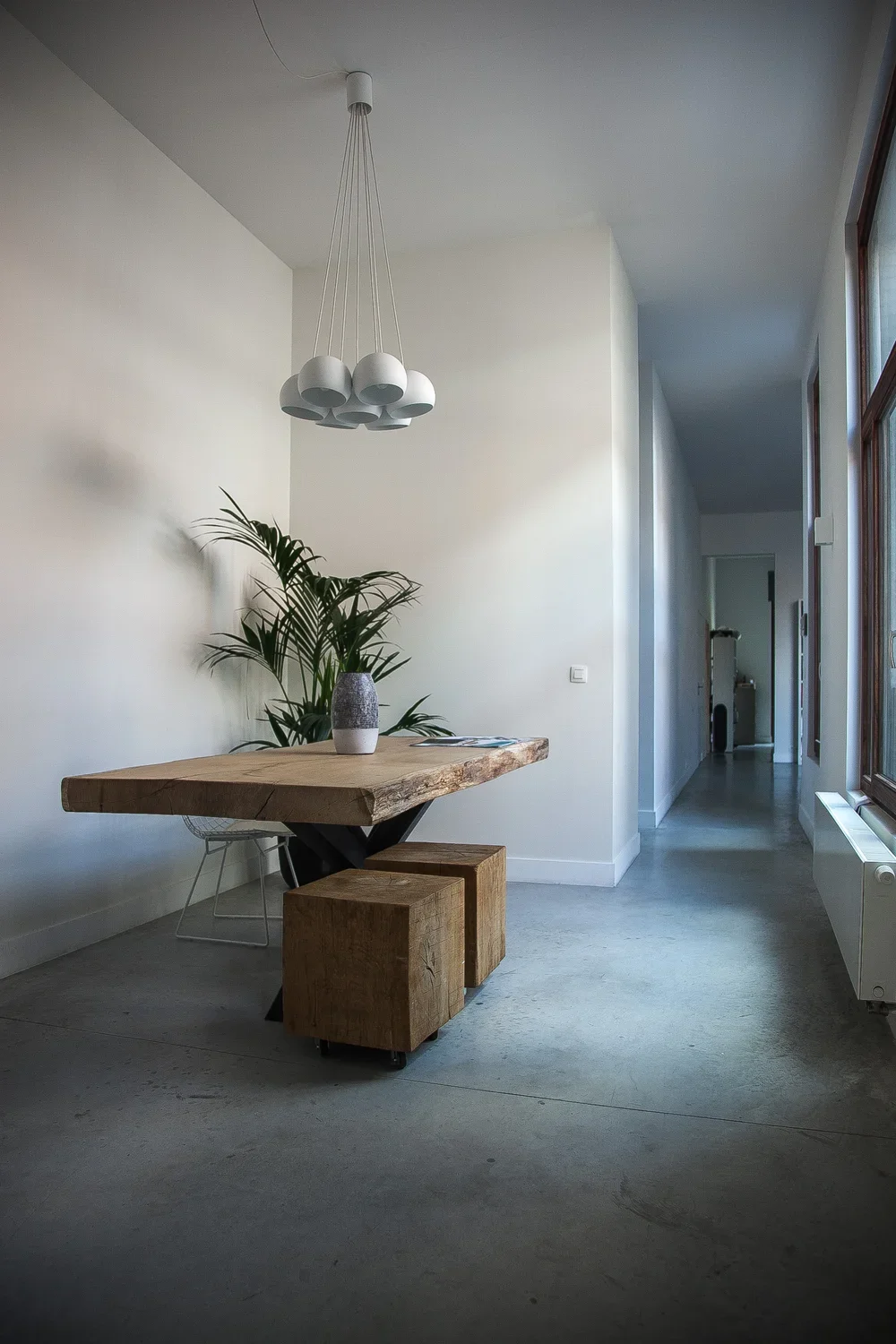 Minimalist dining area with a wooden table, two wooden stools, a vase with a plant, a modern chandelier, and large window on the right letting in natural light.