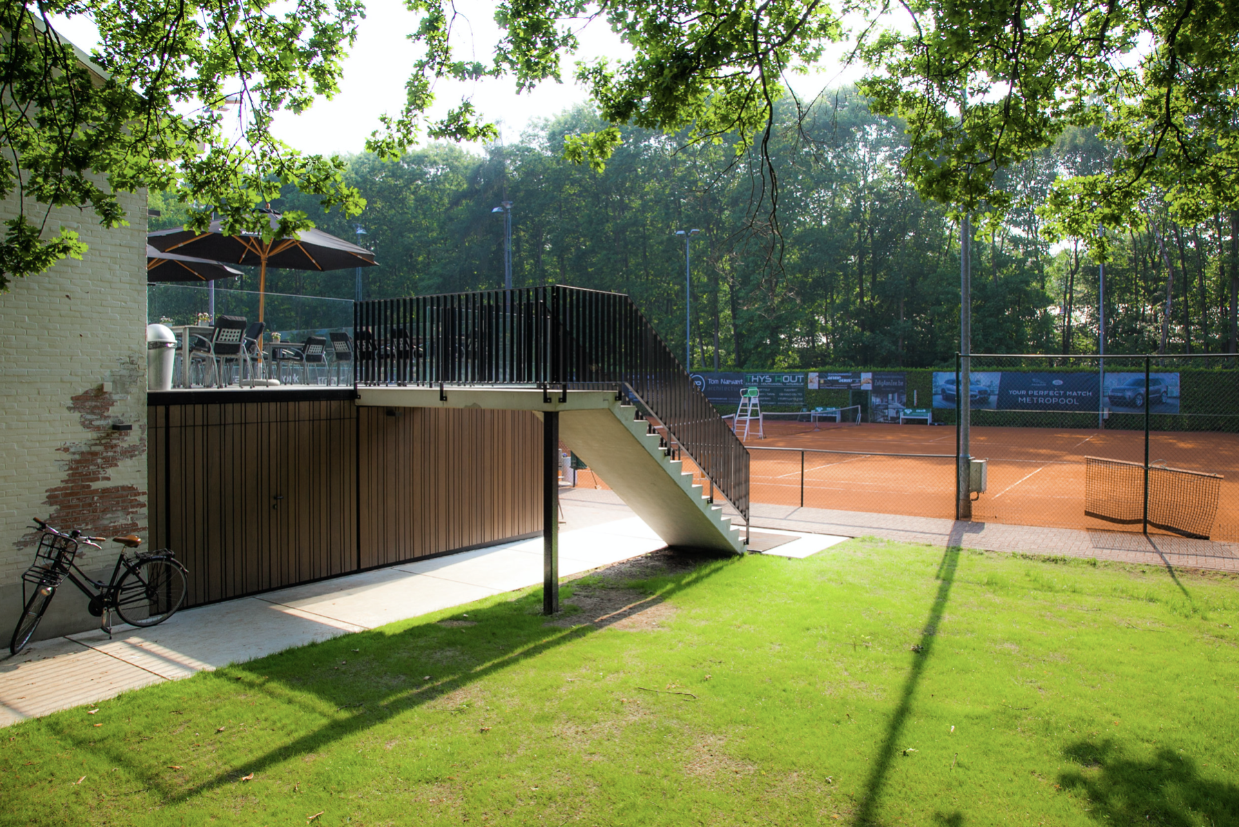 Modern sports clubhouse architecture featuring an elevated viewing deck next to a tennis court, designed by Tom Naeyaert Architecture.