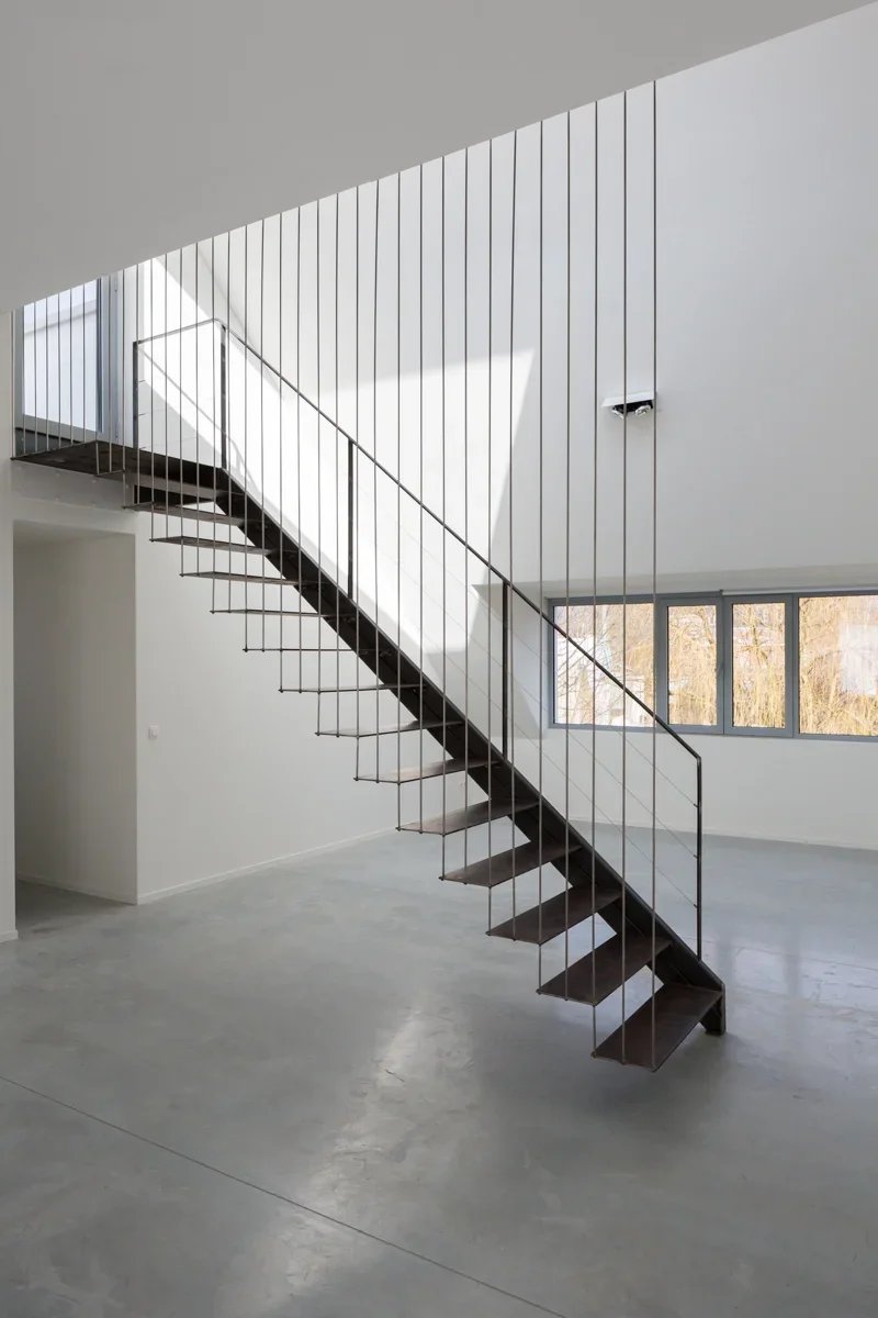 Modern staircase with dark wood steps and metal railing inside a minimalist white room, large windows showing trees outside.