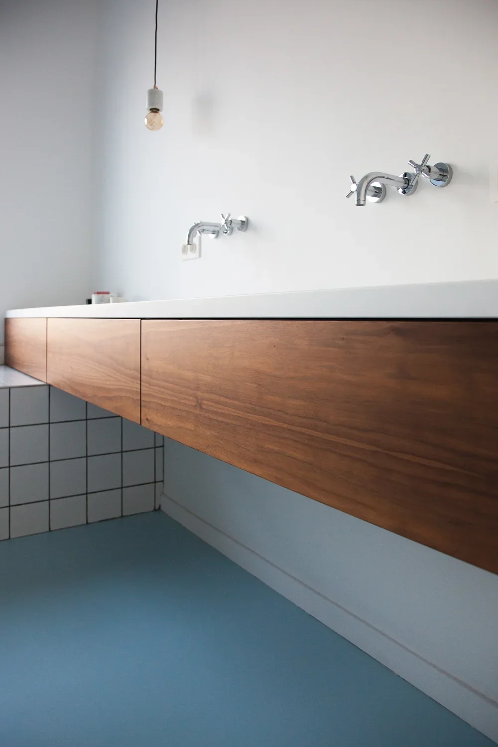 Modern bathroom sink with a white countertop, wooden cabinet, and chrome faucets, with a hanging light bulb and tiled wall.