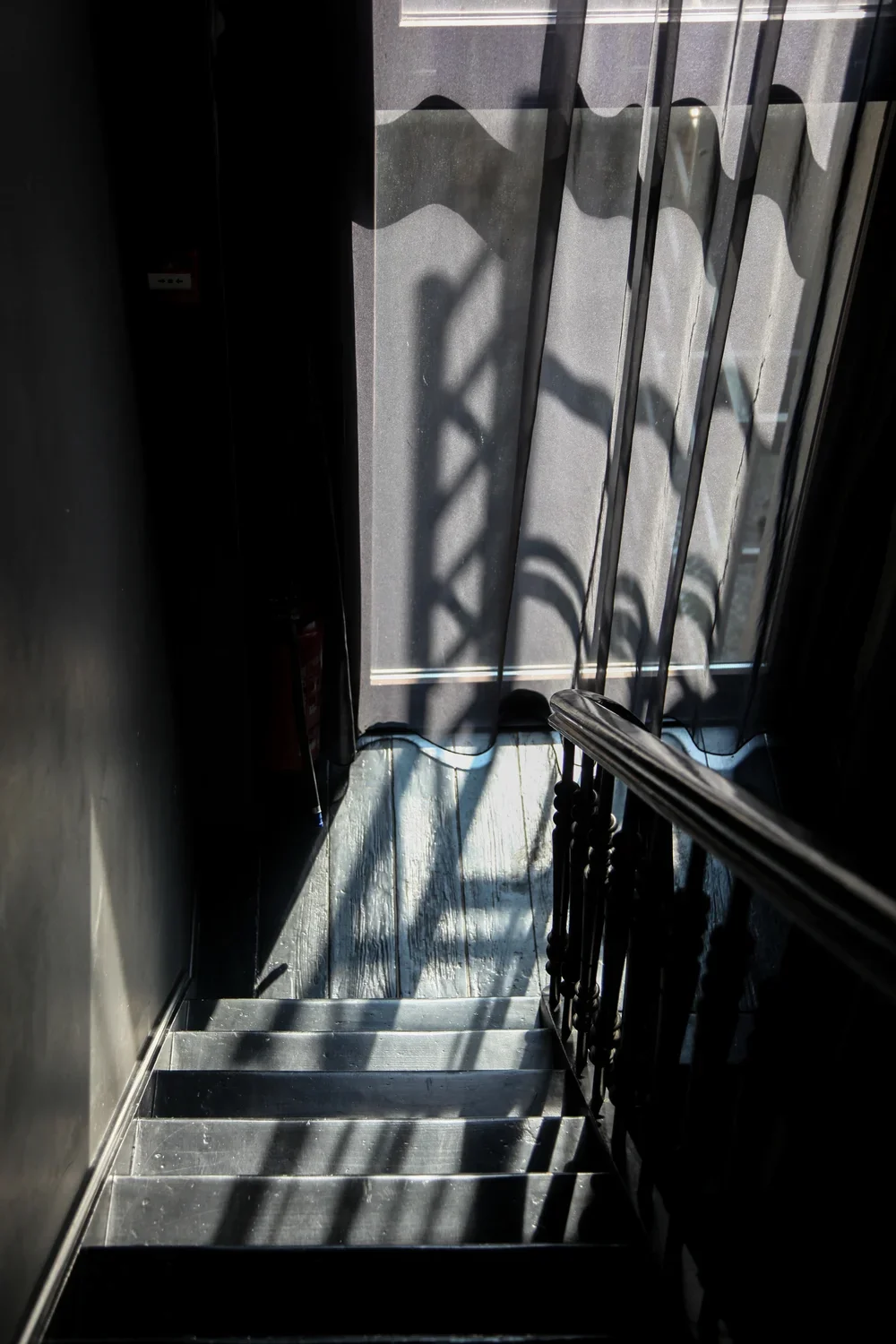 Modern residential staircase leading to an outdoor balcony, featuring dramatic sunlight shadows from a decorative metal railing onto wooden flooring, by Tom Naeyaert Architecture.