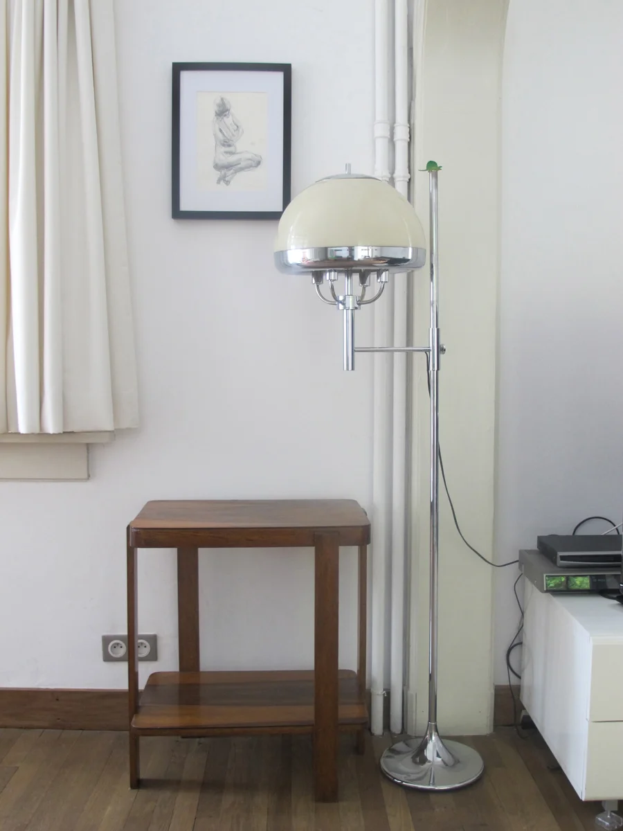 A vintage hair dryer with a large white dome on a tall chrome stand in a room with a wooden side table, framed artwork on the wall, and a white cabinet with electronic devices.