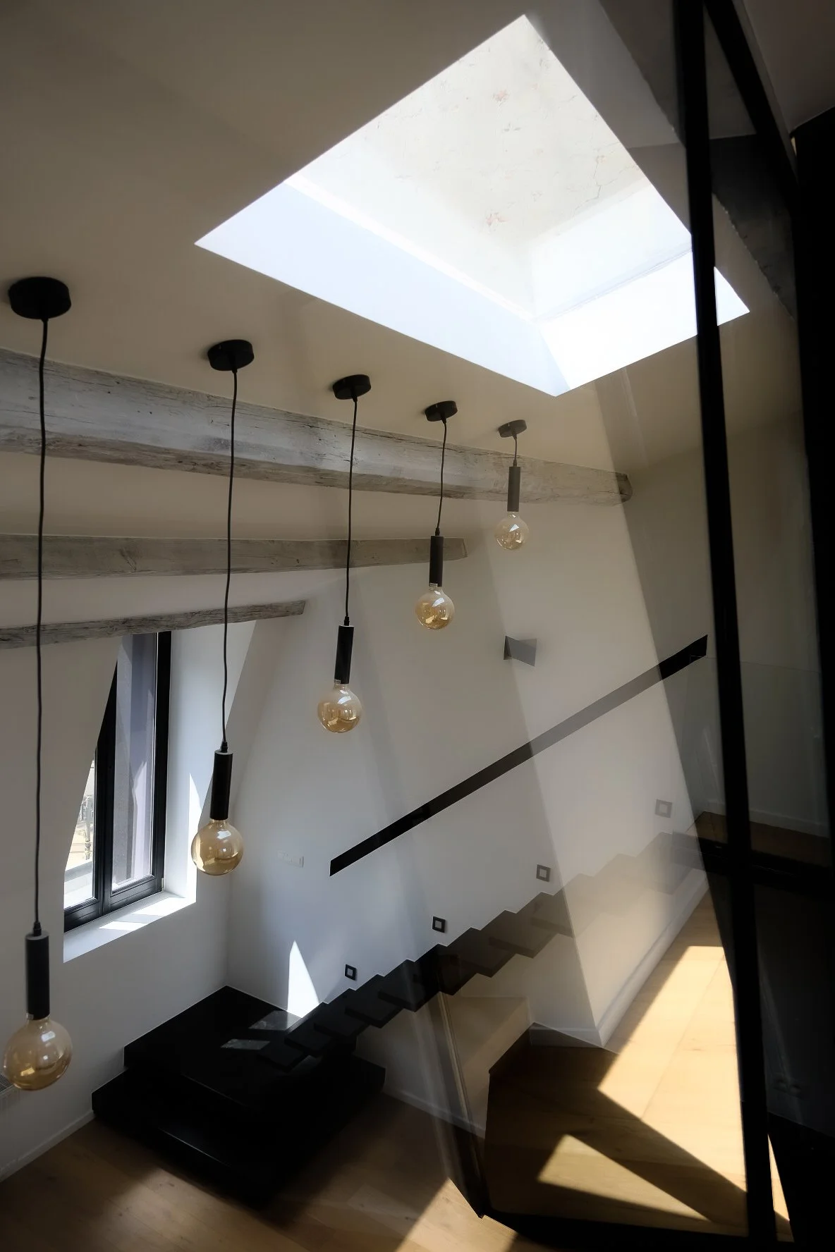 Interior view of a modern staircase featuring hanging pendant lights, a skylight, and exposed wooden beams, designed by Tom Naeyaert Architecture in Antwerp.